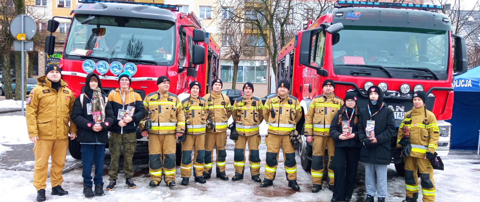 Zdjęcie przedstawia strażaków Komendy Powiatowej Państwowej Straży Pożarnej w Gołdapi, którzy wspólnie z wolontariuszami Wielkiej Orkiestry Świątecznej Pomocy uczestniczą w 32. Finale na Placu Zwycięstwa w Gołdapi. W tle widać dwa pojazdy pożarnicze. Niektóre osoby ujęte na obrazie, posiadają na swojej odzieży naklejone serce Wielkiej Orkiestry Świątecznej Pomocy