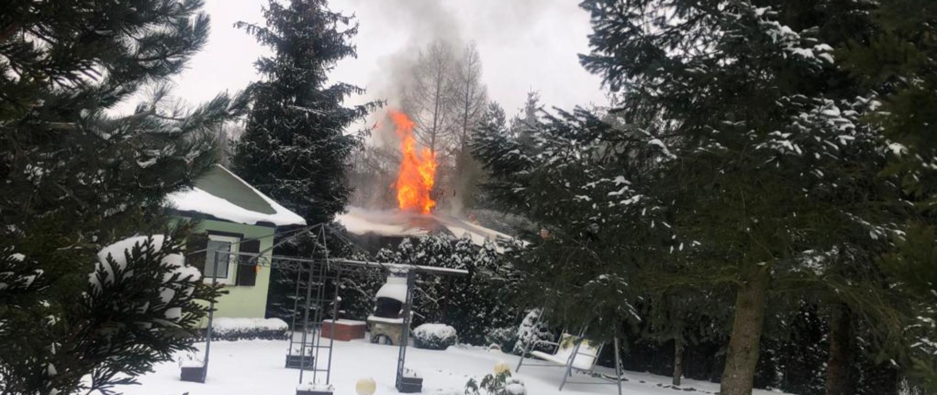 Pora zimowa, zalegający śnieg. Na zdjęciu dużo zieleni oraz drzew iglastych. Po środku zdjęcia języki ognia wychodzące ponad dach budynku. Palący się budynek zasłonięty przez roślinność widać tylko dach.