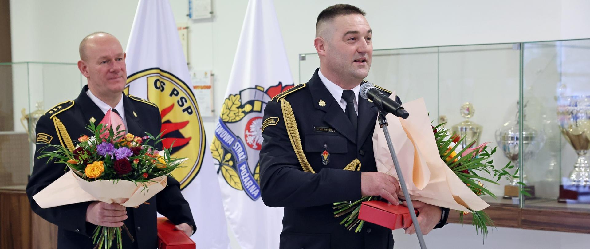 Funkcjonariusz CS PSP z bukietem kwiatów oraz czerwonym etui w dłoni przemawia do mikrofonu, w głębi stojący funkcjonariusz CS PSP trzymający w dłoniach bukiet kwiatów oraz czerwone etui. Obaj w umundurowaniu galowym ze sznurem
