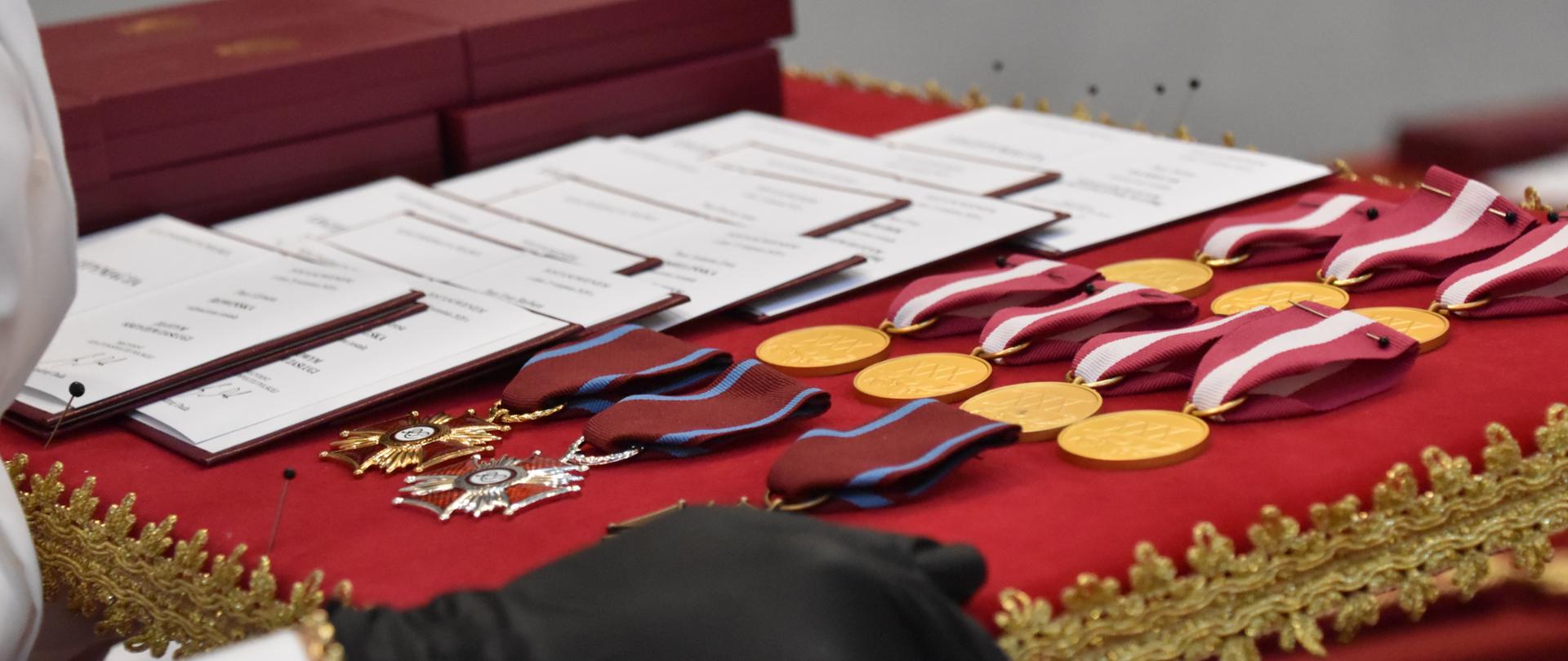 Zdjęcie przedstawia fragment tacy, na której znajdują się odznaczenia państwowe: Krzyże Zasługi oraz Medale za Długoletnią Służbę oraz decyzje Prezydenta o przyznaniu tych wyróżnień. Tacę tę trzyma w ręku kobieta asystująca.