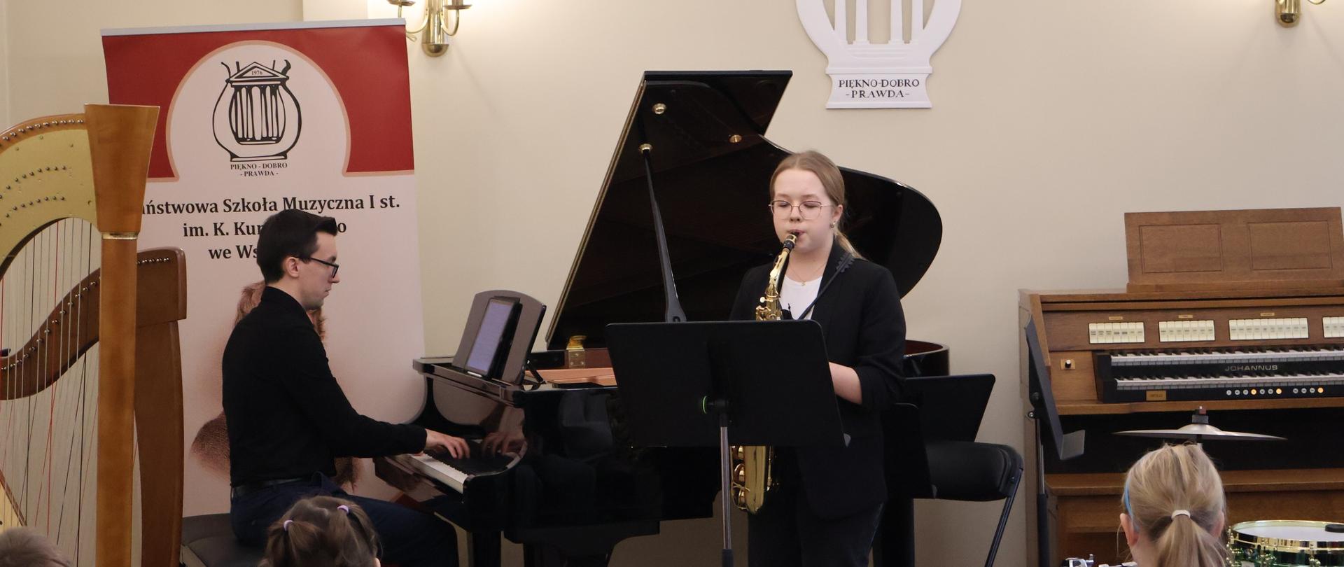 Nastolatka grająca na saksofonie przed publicznością przedszkolaków. Przy fortepianie nauczyciel.