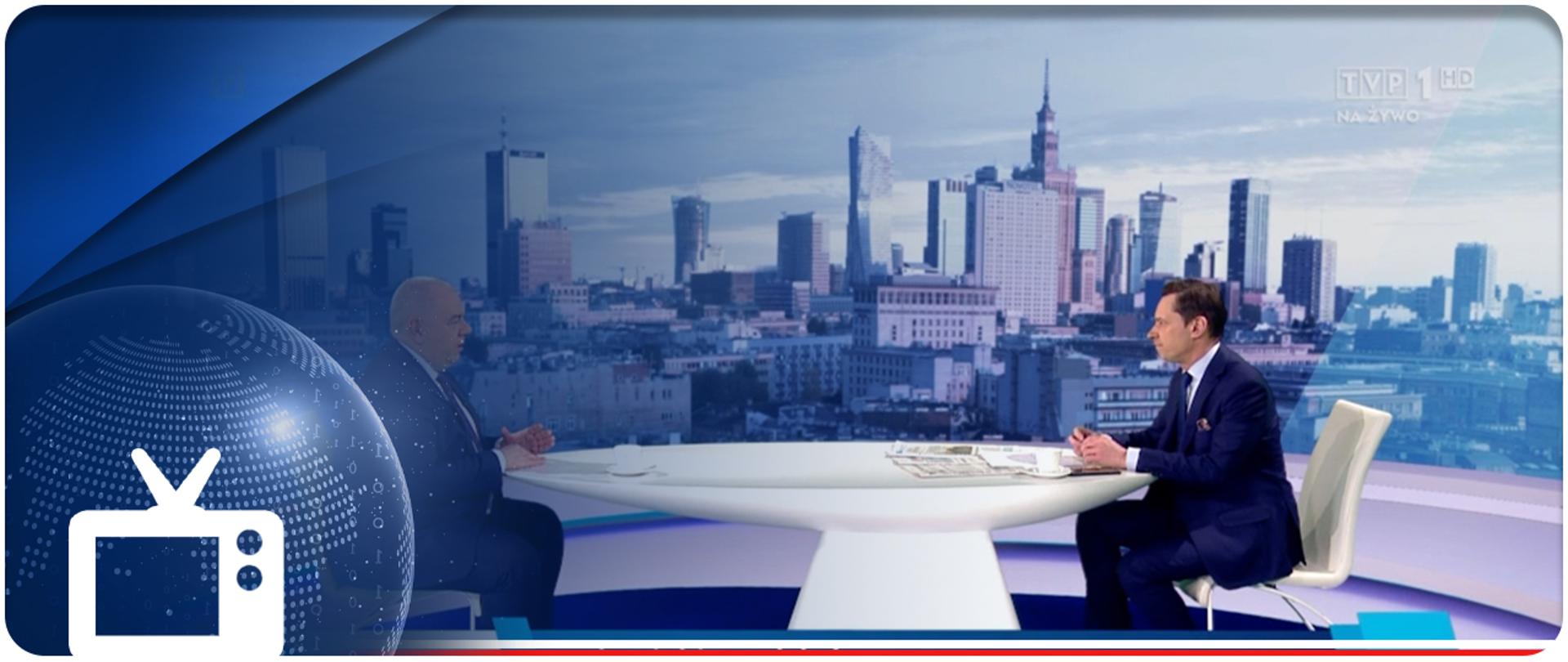 Kadr z programu publicystycznego: przy białym stole siedzą (po lewej) wicepremier Jacek Sasin, (po prawej) prowadzący Krzysztof Ziemiec. W tle panorama Warszawy. Na obrazek nałożona granatowa apla i piktogram odbiornika telewizyjnego
