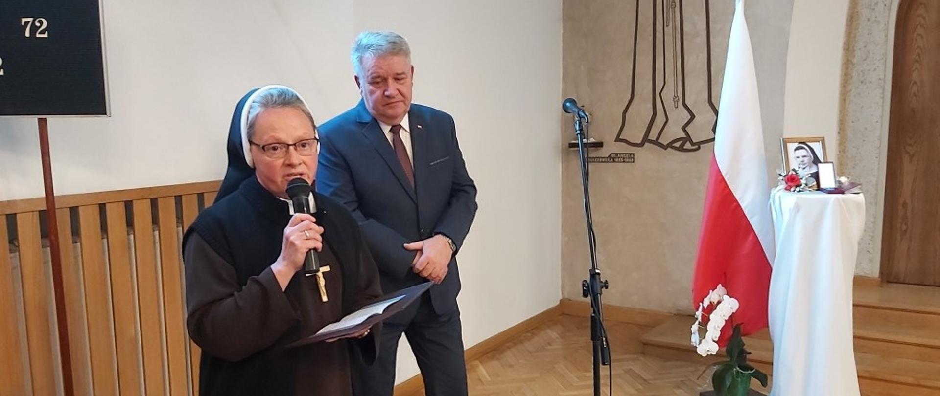 Na zdjęciu od lewej Siostra Marii Christelli Skwarek w momencie przemówienia. Na drugim planie Wicewojewoda Sylwester Dąbrowski. W głębi stoi wysoki stojak przykryty białym materiałem, na którym znajduje się zdjęcie z wizerunkiem uhonorowanej śp. Matki Marii Anzelmy Trojanowskiej. Przy stole polska flaga.