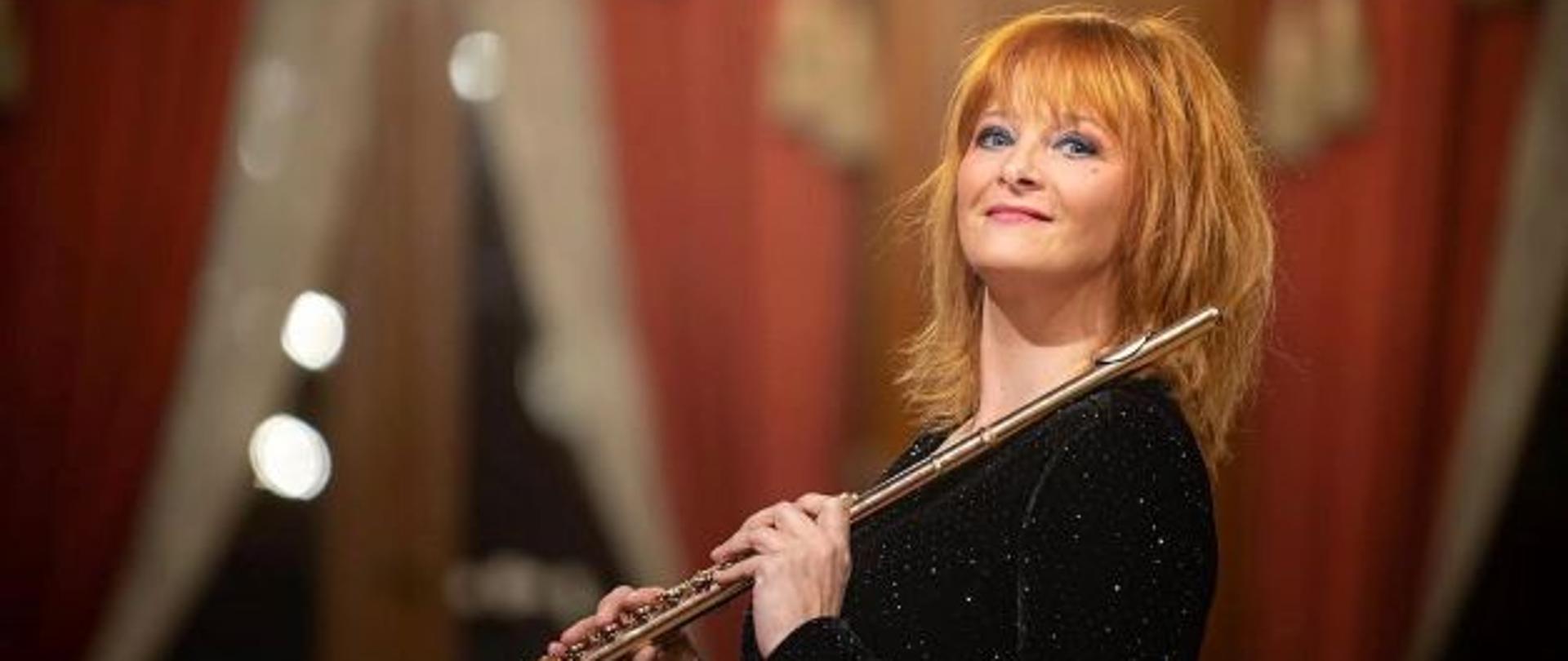 Zdjęcie przedstawia prof. dr hab. Ewelinę Zawiślak, która trzyma flet poprzeczny w tle scena z kotarą.