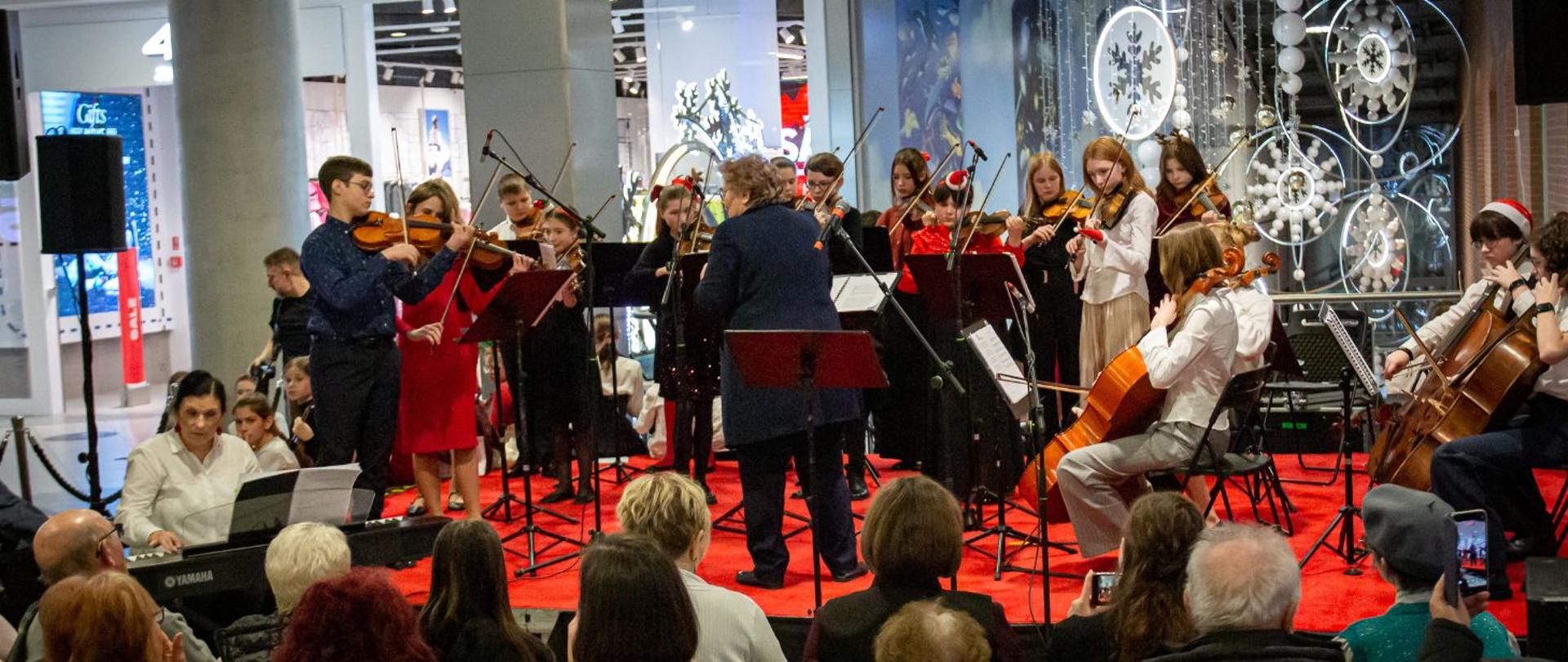Na zdjęciu widać grupę młodych muzyków występujących przed publicznością w galerii handlowej Focus Park Rybnik. Na scenie, pokrytej czerwonym dywanem, znajduje się zespół instrumentalny złożony głównie ze skrzypków, wiolonczelistów i pianisty. Muzycy są elegancko ubrani — część z nich ma stroje w kolorach białym, czarnym i czerwonym.
Na środku stoi dyrygent, zwrócony plecami do publiczności, prowadzący zespół. W tle widać dekoracje zimowe — duże śnieżynki i błyszczące ozdoby. Przed sceną siedzi publiczność, uważnie słuchająca występu. Cała scena jest dobrze oświetlona i panuje przyjazna, świąteczna atmosfera.