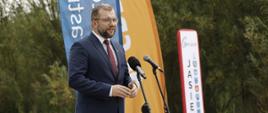 Minister funduszy i polityki regionalnej Grzegorz Puda stoi przed dwoma mikrofonami, za jego plecami tablica z napisem "Jasienica" i windery GDDKiA i Min. Infrastruktury