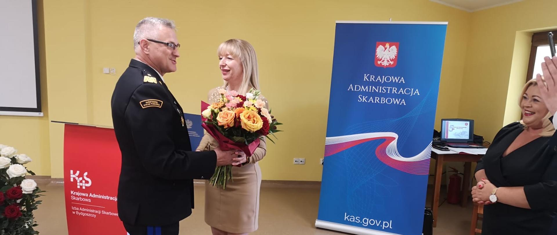 Z lewej Kujawsko-Pomorski Komendant Wojewódzki, obok niego Dyrektor. KAS. Komendant w granatowym mundurze wyjściowym wręcza Pani Dyrektor bukiet kwiatów. Po lewej w tle baner z napisem Krajowa Administracja Skarbowa.