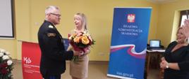 Z lewej Kujawsko-Pomorski Komendant Wojewódzki, obok niego Dyrektor. KAS. Komendant w granatowym mundurze wyjściowym wręcza Pani Dyrektor bukiet kwiatów. Po lewej w tle baner z napisem Krajowa Administracja Skarbowa.