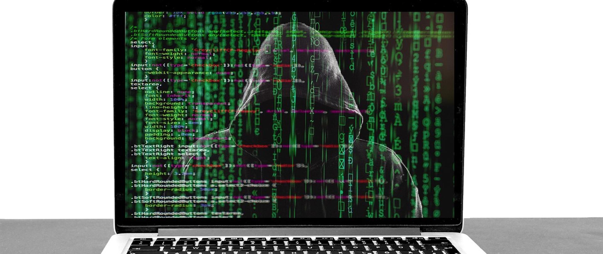 laptop, na ekranie laptopa widać postać w kapturze i dużo zielonych cyferek i literek porozrzucanych na całym ekranie