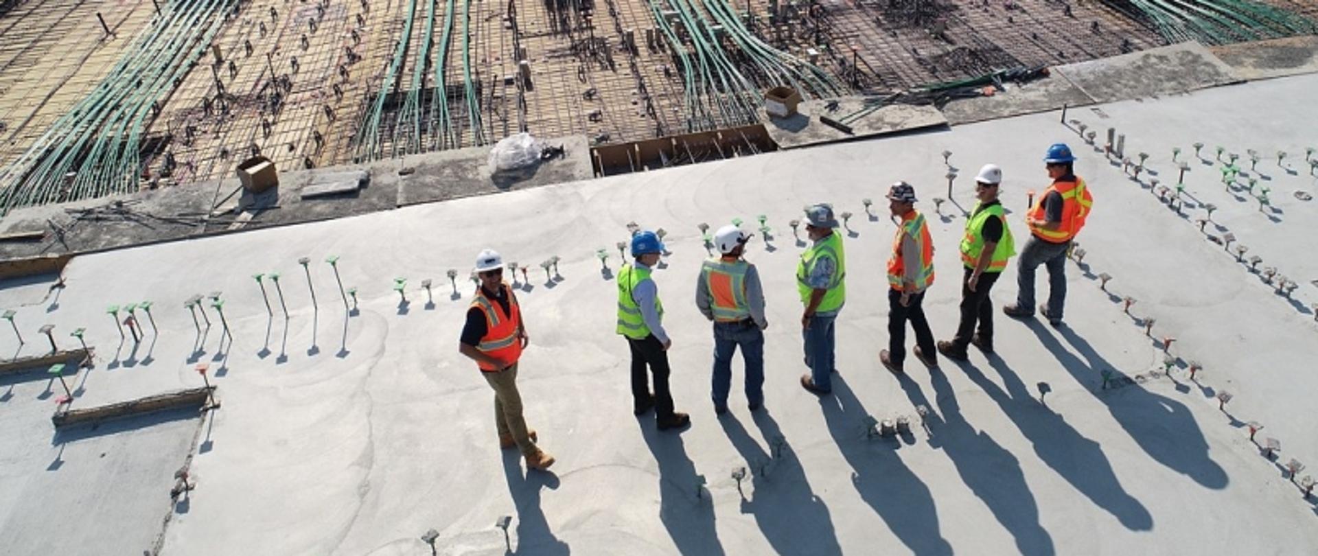 Siedmiu pracowników budowy w kaskach i kamizelkach na tle materiałów budowlanych, stojących na betonowej powierzchni.
