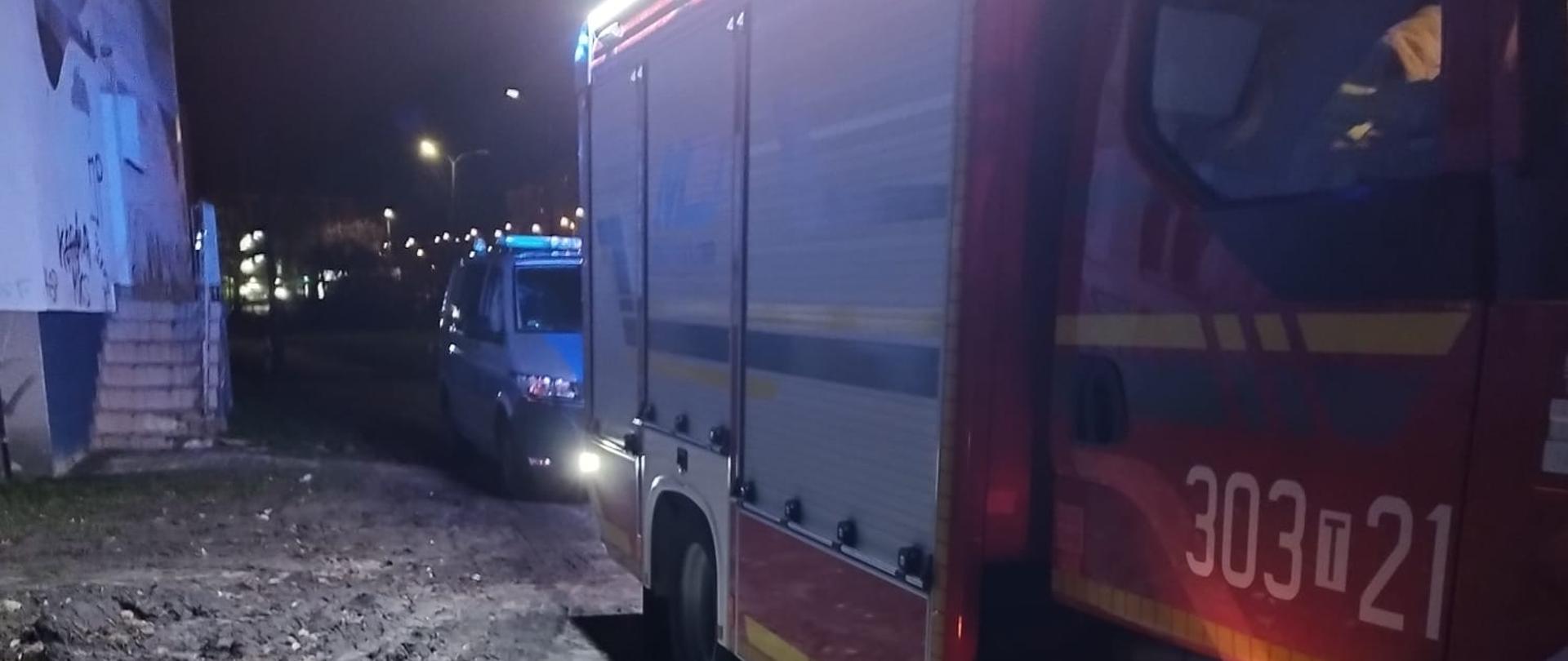 Zdjęcie przedstawia samochód pożarniczy ustawiony przy budynku wielorodzinnym (po lewej stronie) gdzie doszło do pożaru tapicerowanego mebla.