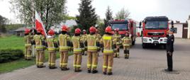 Zdjęcie obrazuje moment zmiany służbowej w komendzie w Aleksandrowie Kujawskim podczas uroczystej zbiórki z okazji Dnia Flagi. w tle samochody strażackie