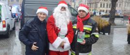 Korowód Wielkich Serc. Rawicki rynek . Na pierwszym planie, w towarzystwie św. Mikołaja do wspólnego zdjęcia pozują: policjantka i strażak ubrani w elementu stroju świętego Mikołaja. Strażak w lewej ręce trzyma specjalną skarbonkę. W tle drzewo, samochody oraz kamienice.