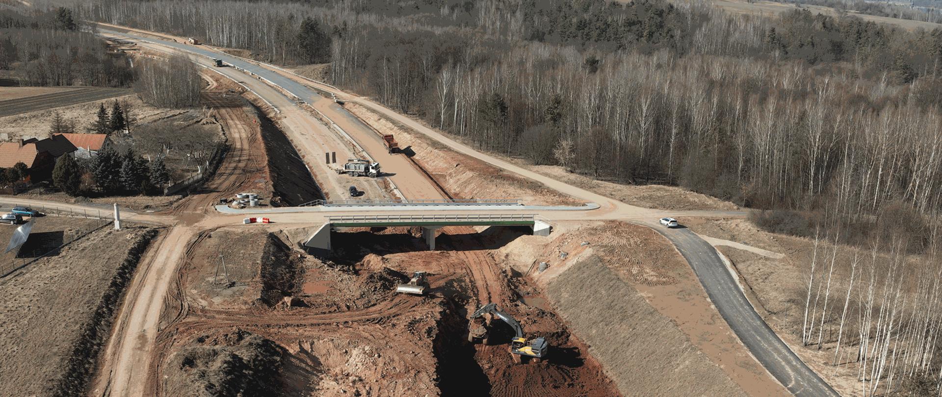 Budowa obwodnicy Wąchocka - roboty ziemne i nawierzchniowe w śladzie dwujezdniowej trasy. Na pierwszym planie wykop, wiadukt nad wykopem, za wiaduktem jezdnie z warstwą nawierzchni. W wykopie spycharka i koparka. Zdjęcie wykonane z drona. Po prawej stronie drogi las. 