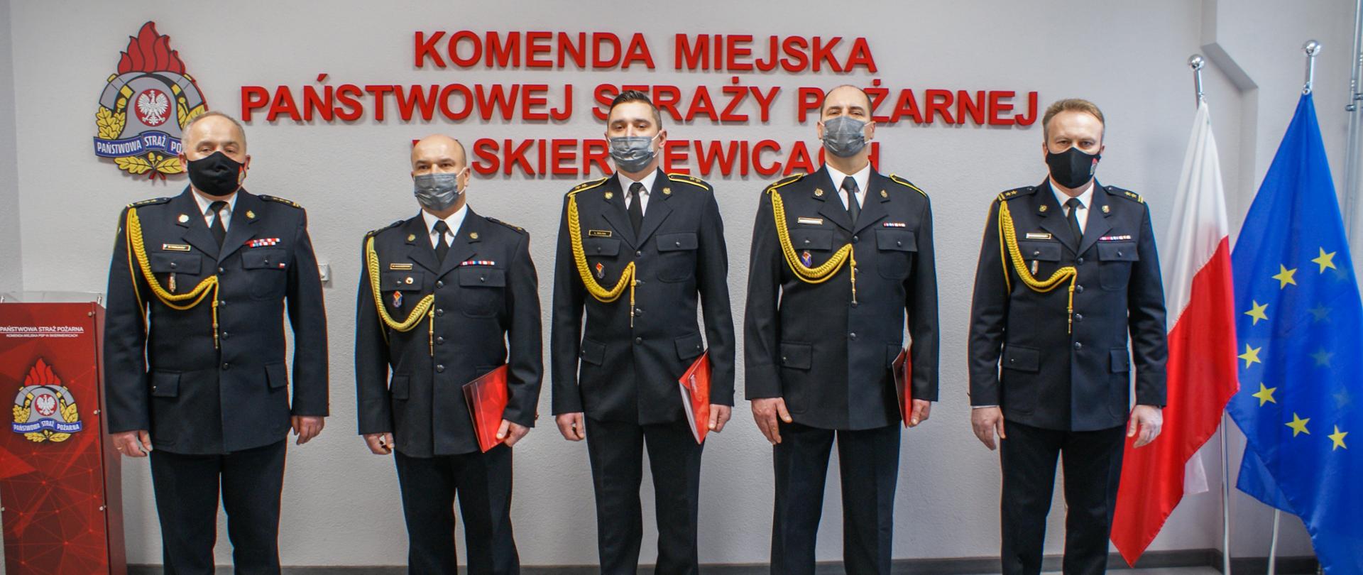 Zdjęcie przedstawia pięciu strażaków komendy miejskiej w mundurach wyjściowych koloru czarnego na tle czerwonego napisu Komenda Miejska Państwowej Straży Pożarnej w Skierniewicach, loga psp, flag polski i unii europejskiej oraz godła państwowego. 