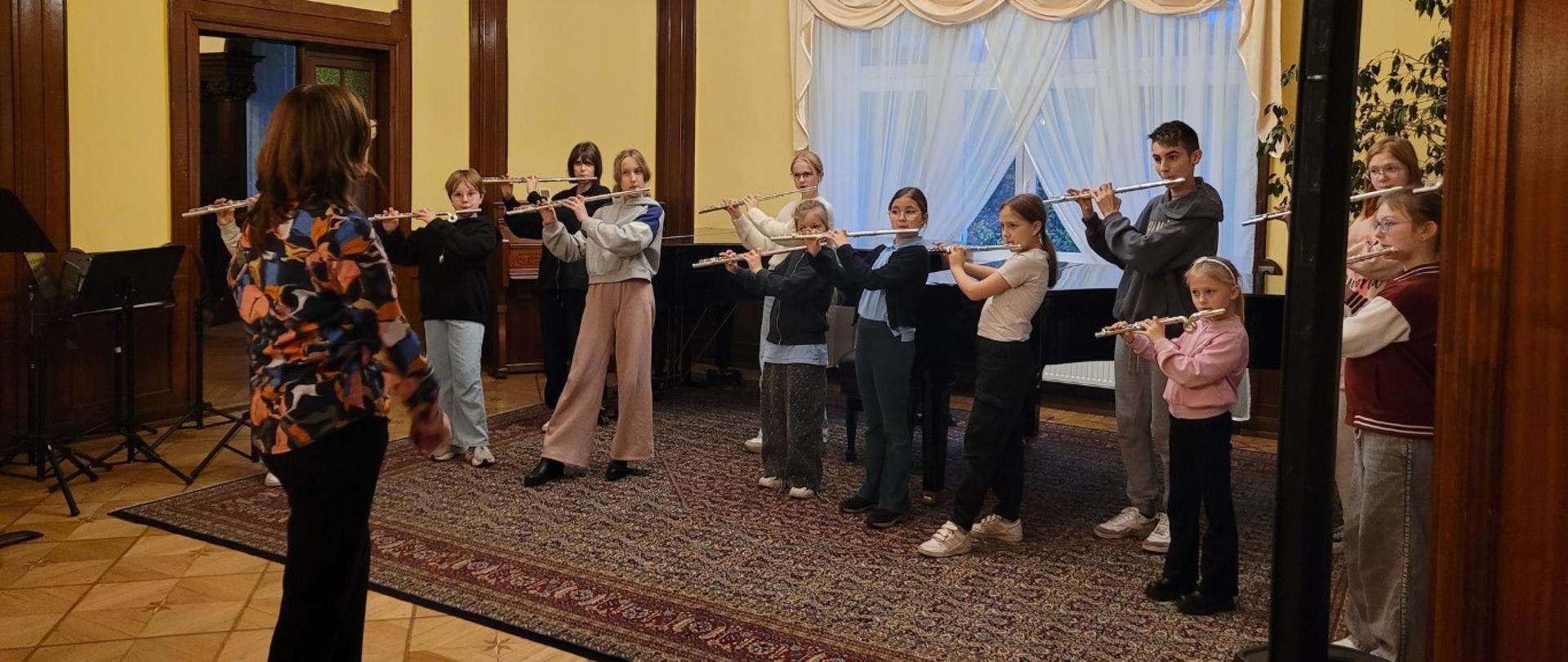 Zdjęcie kolorowe, dziesięcioro dzieci i młodzieży stoi w rzędzie grających na fletach poprzecznych, z lewej tyłem stoi kobieta w kolorowej bluzce, instruktorka, sala z jasnymi, drewnianymi panelami na ścianach, po prawej stronie czarna kolumna i roślina w doniczce, z tyłu okna z beżowymi zasłonami.