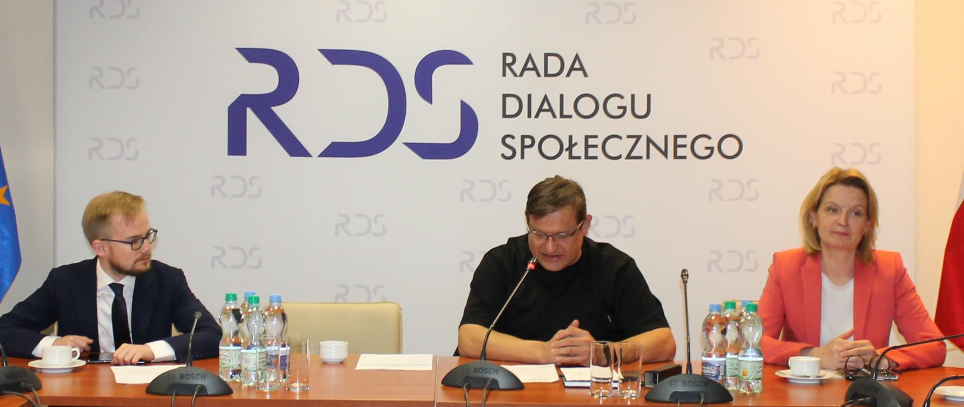 Na zdjęciu widnieje stół prezydialny za którym siedzą następujące osoby od lewej strony: Piotr Patkowski - Podsekretarz Stanu w Ministerstwie Finansów, Jacek Męcina - Przewodniczący Zespołu ds. budżetu, wynagrodzeń i świadczeń socjalnych oraz Barbara Socha - Podsekretarz Stanu w Ministerstwie Rodziny i Polityki Społecznej. W tle siedzących osób widnieje baner z napisem Rada Dialogu Społecznego. Każda z osób ma przed sobą mikrofon.