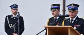 Dwóch strażaków stoi przy mównicy. Strażak z prawej strony przemawia przez mikrofon. Mikrofon jest koloru czarnego. Strażacy mają ubrany mundur wyjściowy koloru czarnego. Strażacy mają ubrane rogatywki z godłem Polski, przypięte do munduru odznaczenia oraz złoty sznur galowy. Z lewej strony przy drugim mikrofonie stoi strażak ubrany w mundur wyjściowy koloru czarnego. Strażak ma ubraną rogatywkę, ma przypięte odznaczenia do munduru i sznur galowy koloru srebrnego. W tle widać białą ścianę budynku. 