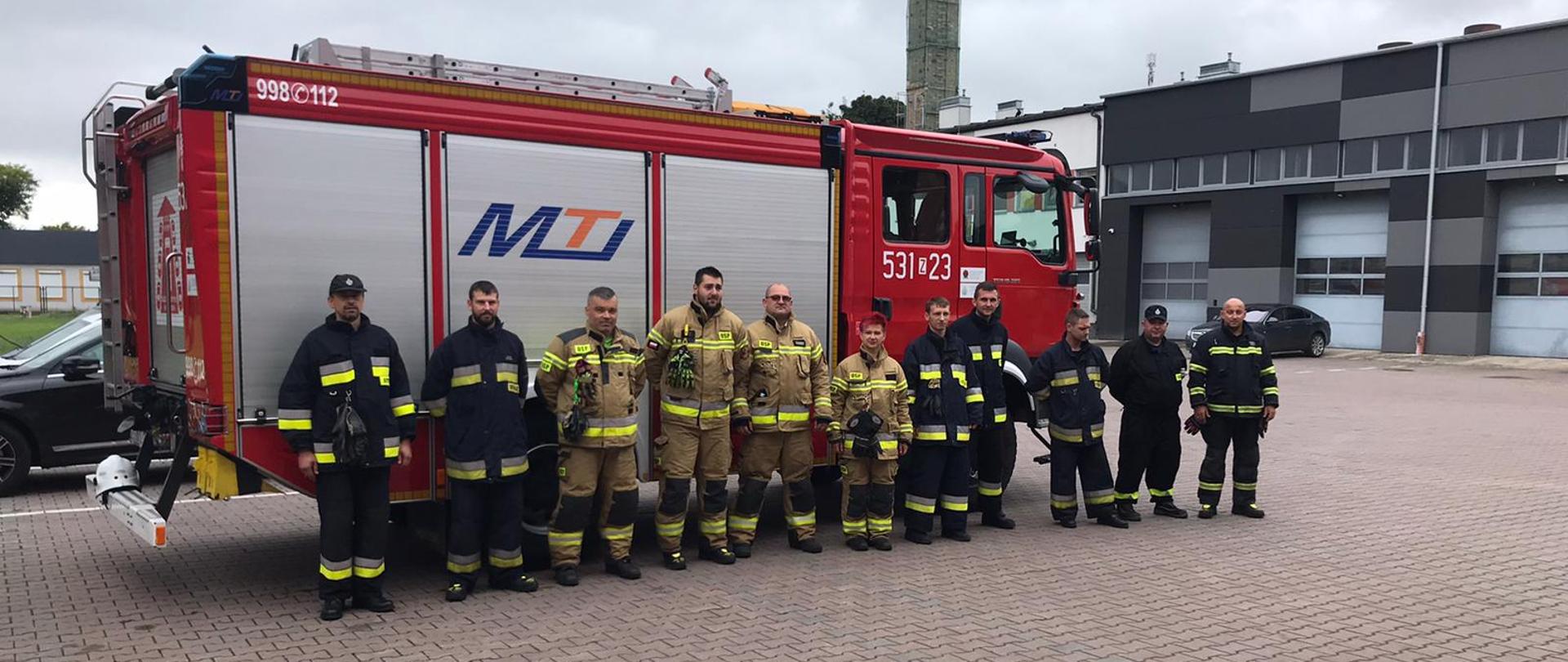 Grupa strażaków w umundurowaniu specjalnym na tle samochodu pożarniczego 