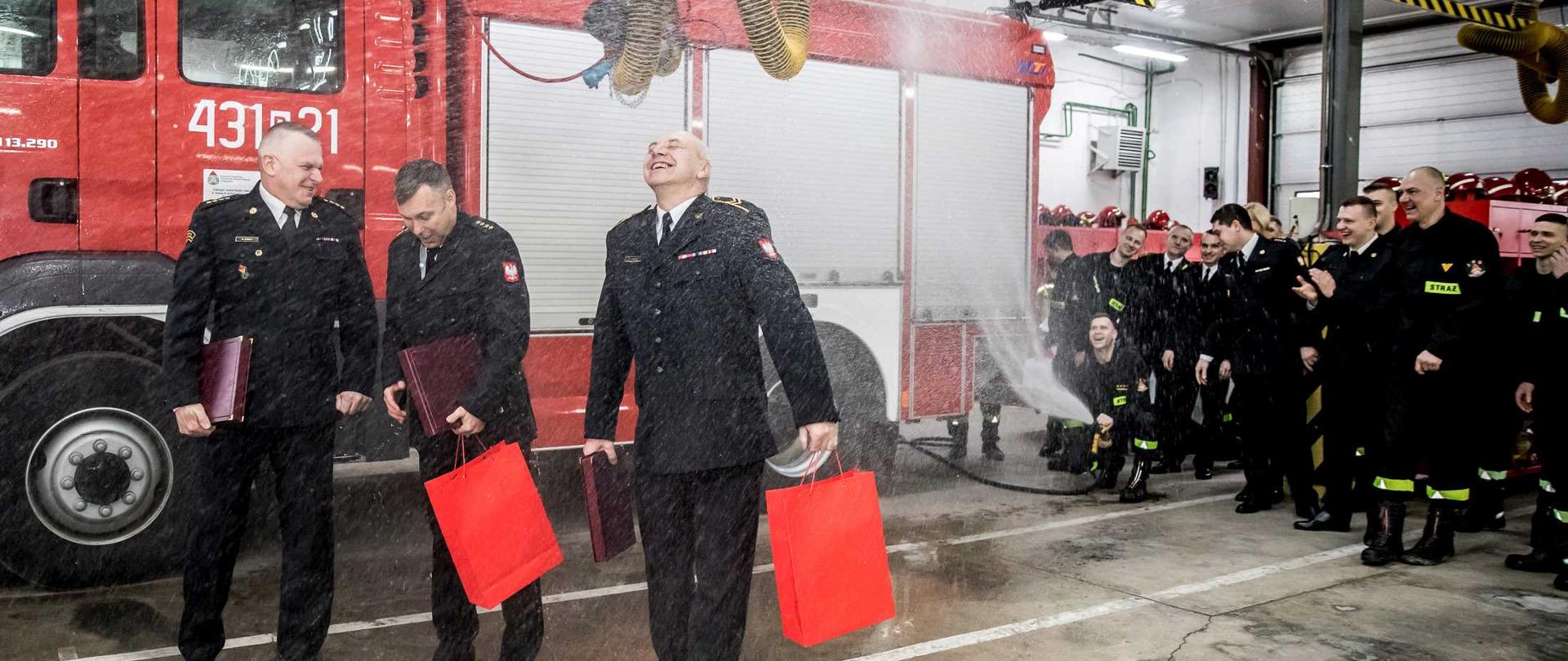 Na tle czerwonego samochodu stoi trzech strażaków, którzy polewani są wodą. Z tyłu stoją inni strażacy.