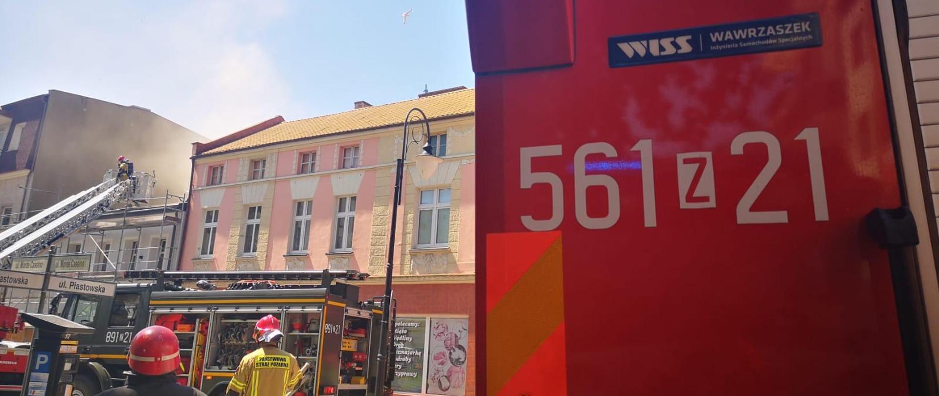 Zdjęcie przedstawia budynek mieszkalny wielorodzinny w zwartej zabudowie. Widoczni strażacy podczas działań. Po prawej stronie duży napis na samochodzie pożarniczym koloru czerwonego 561z21.