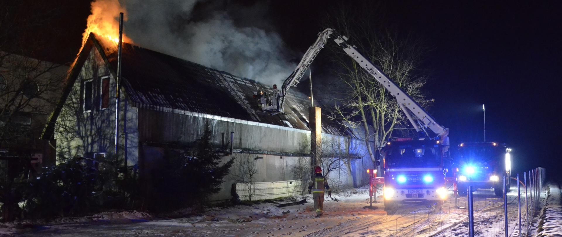 Na zdjęciu widać dwa samochody ratowniczo gaśnicze, stojący przy palącym się budynku. Jeden z nich to drabina z koszem w którym stoi strażak gaszący pożar natomiast drugi to samochód z wodą.