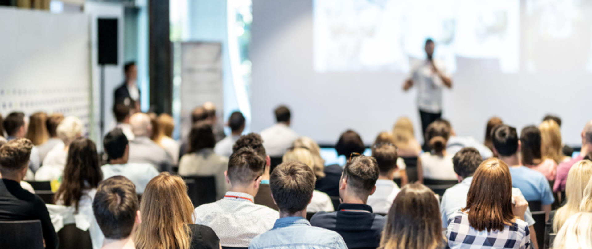 Widok z tyłu na publiczność podczas wystąpienia w sali konferencyjnej; prelegent stoi przed dużym ekranem.
