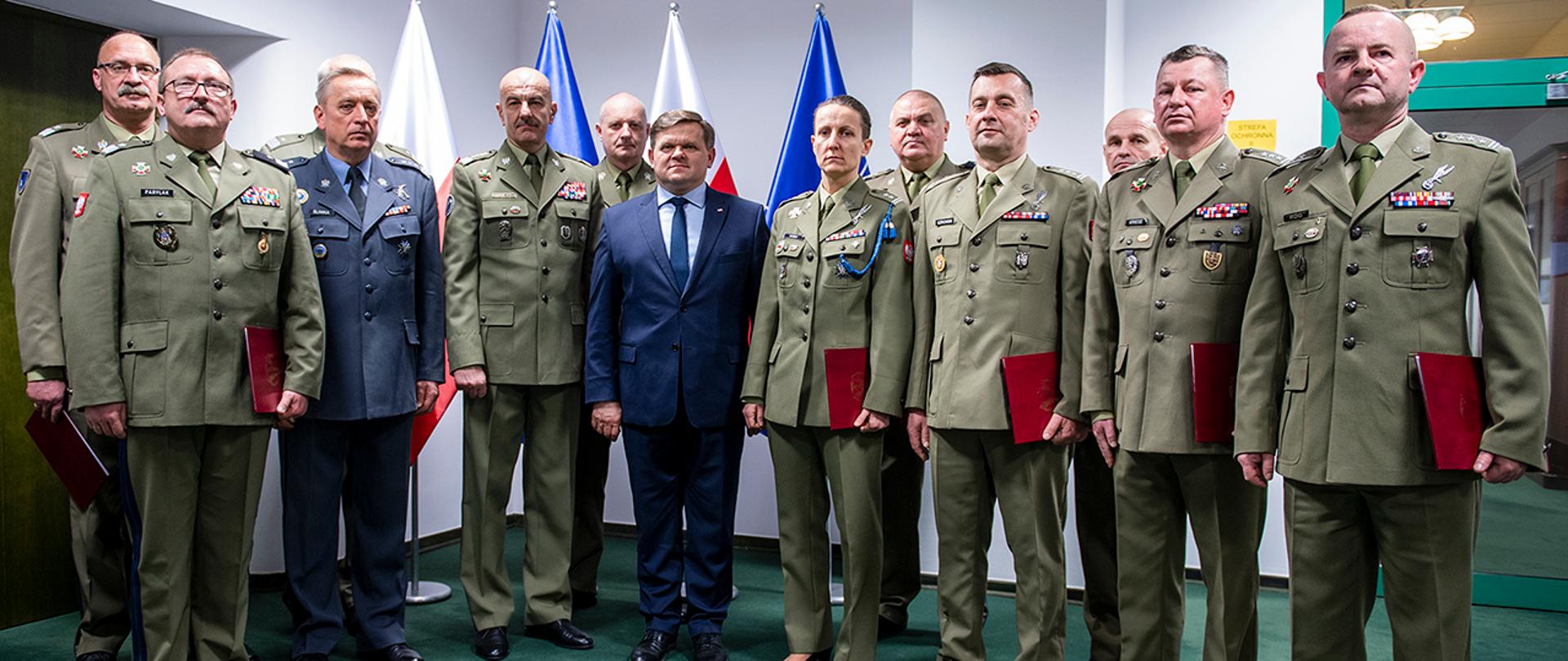 Wojciech Skurkiewicz, sekretarz stanu w MON stoi z nowo mianowanymi żołnierzami Wojska Polskiego. W tle flagi Polski i NATO.