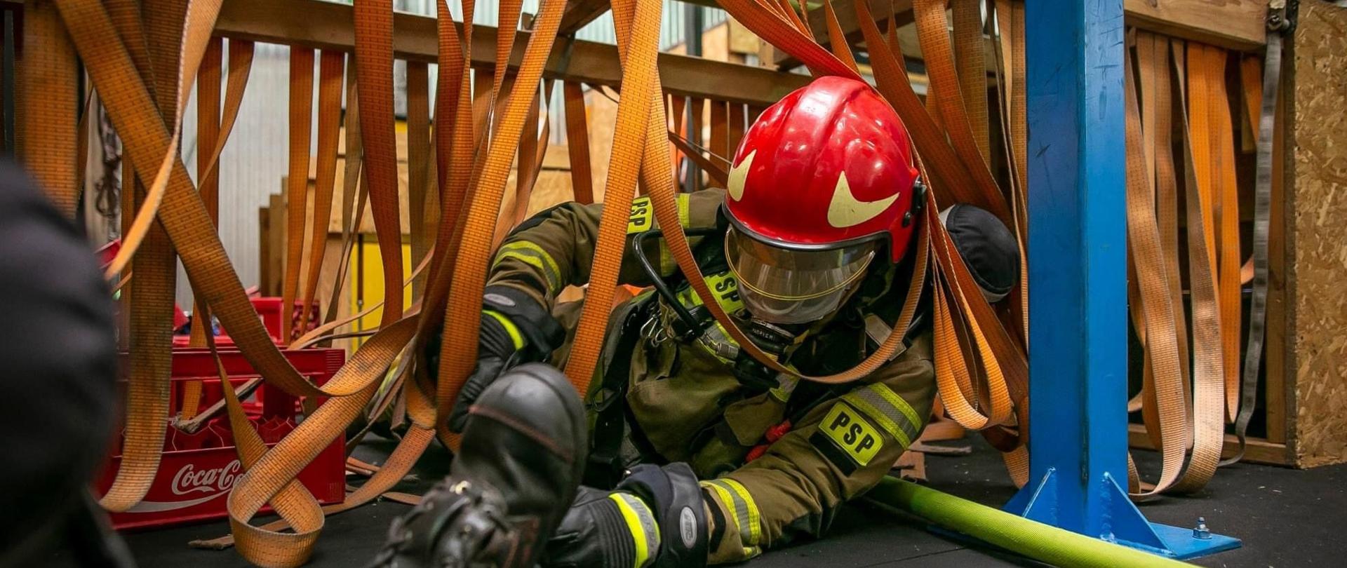 Strażak w umundurowaniu specjalnym przechodzący z wężem przez drewniany tor przeszkód z podwieszonymi pasami transportowymi.