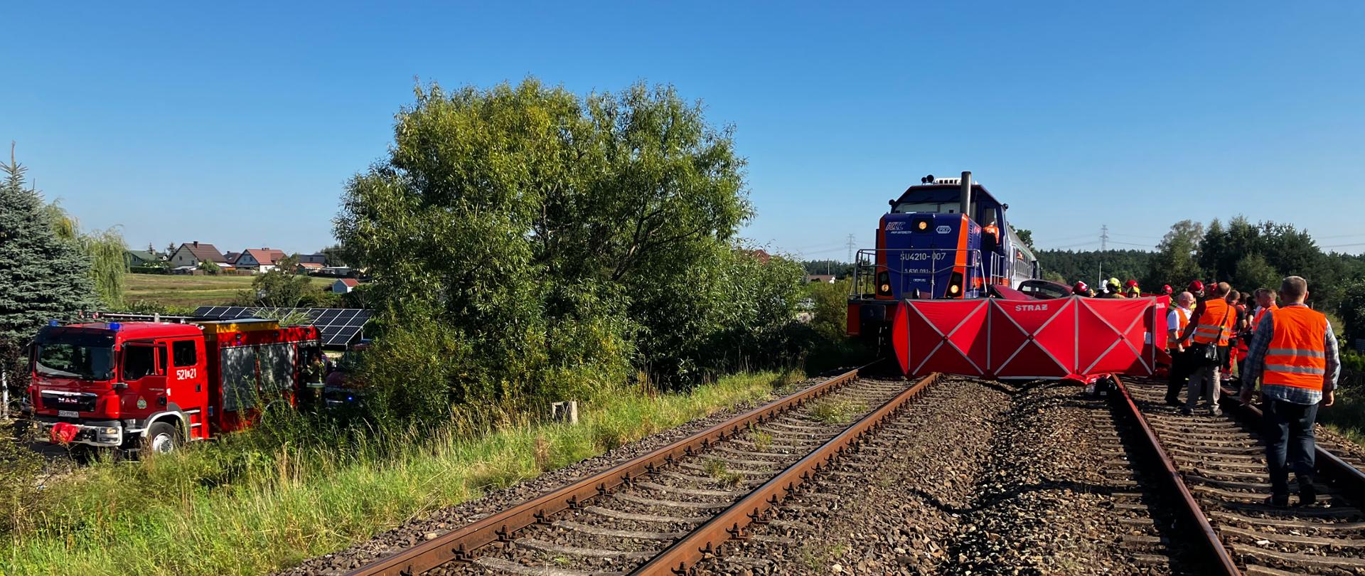 Na zdjęciu widać miejsce zdarzenia - torowisko i lokomotywę. Z przodu parawan straży pożarnej. w lewej części zdjęcia samochód pożarniczy, wiele osób ze służb ratowniczych i kolei.