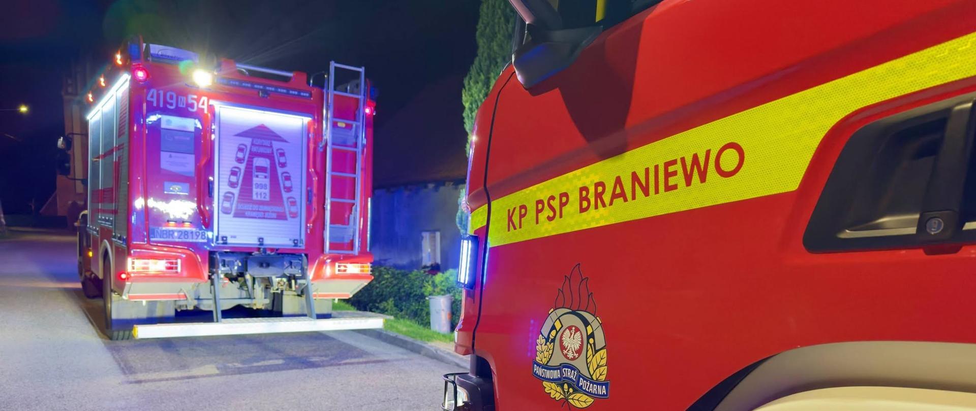 Na zdjęciu widoczne są dwa samochody ratowniczo-gaśnicze Komendy Powiatowej PSP w Braniewie podczas nocnych działań. Pojazdy mają włączone oświetlenie oraz sygnały uprzywilejowania, zabezpieczając miejsce prowadzonej akcji. Zdjęcie przedstawia zaplecze sprzętowe strażaków wykorzystywane podczas działań ratowniczych.