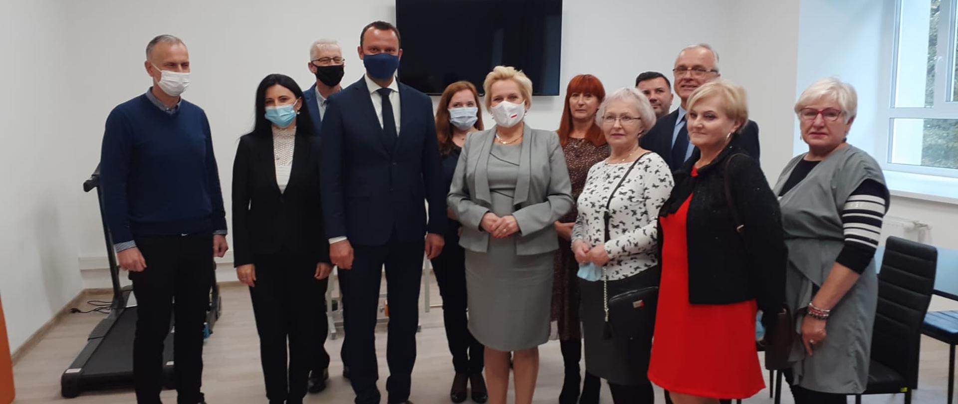 Na zdjęciu uczestnicy uroczystego otwarcia placówki dla osób starszych w miejscowości Paczków. Zdjęcie prezentuje grupę ludzi, stojących koło siebie pozujących do zdjęcia. W środku stoi wicewojewoda opolski. Wszyscy ubrani są elegancko. W tle widać sprzęt sportowy, zakupiony ze środków budżetu państwa, jest to m.in. bieżnia. Na ścianie za pozującymi wisi telewizor. Po prawej stronie kadru w tle widać czarne krzesła. 