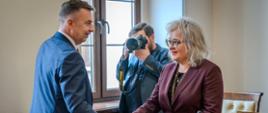 Minister Wieczorek stoi i podaje rękę kobiecie w fioletowej marynarce, za nimi mężczyzna robi im zdjęcie aparatem.