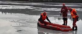 Trzech strażaków w specjalistycznym umundurowaniu razem z saniami lodowymi stoi na zamarzniętym jeziorze.