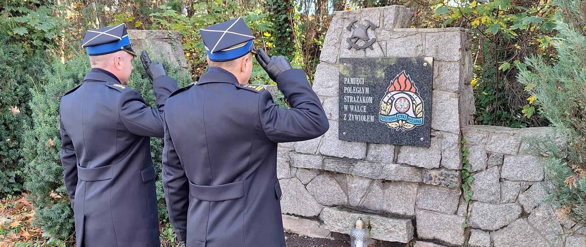 Komendanci zapalili znicz przy tablicy poświęconej strażakom. Komendanci stoją przodem do tablicy i oddają honor. 