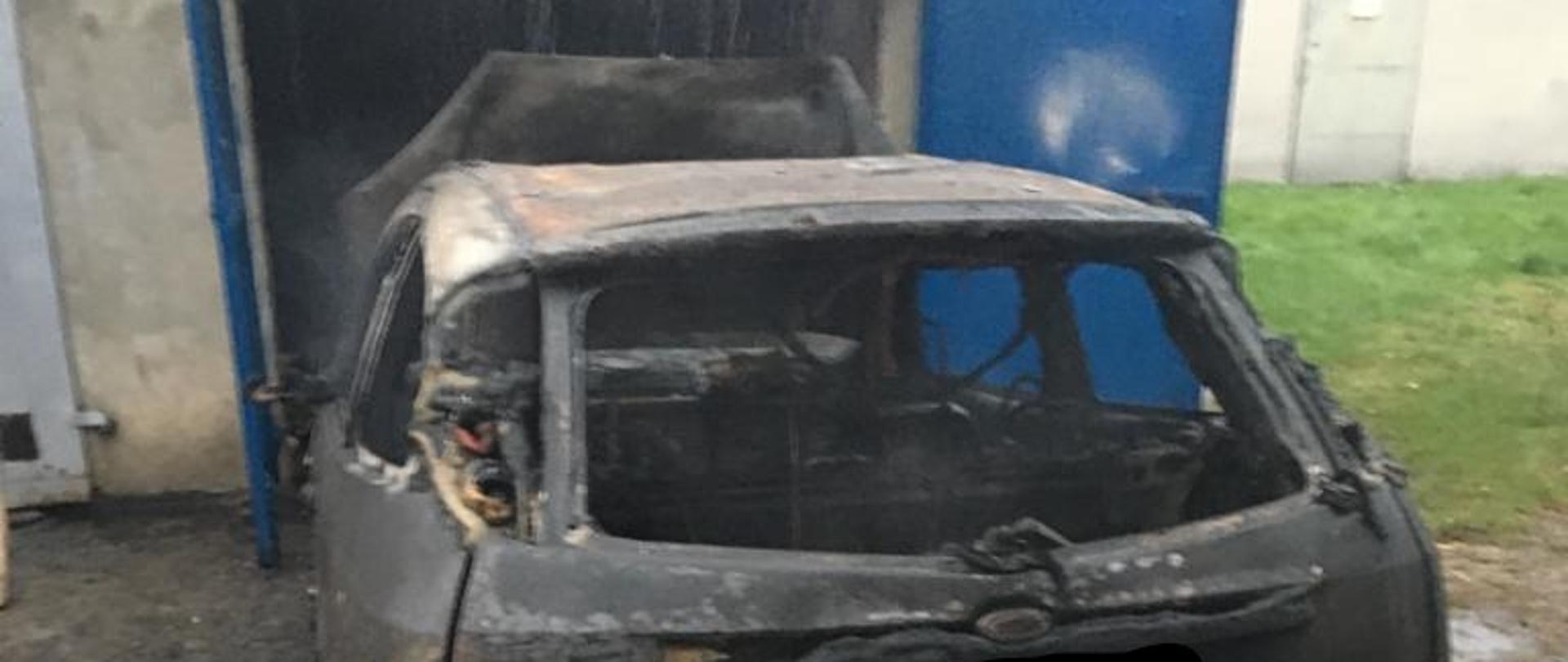 Centralnie na zdjęciu znajduje się samochód osobowy, który w wyniku pożaru uległ całkowicie zniszczeniu. Również widać murowany garaż w którym powstał pożar.