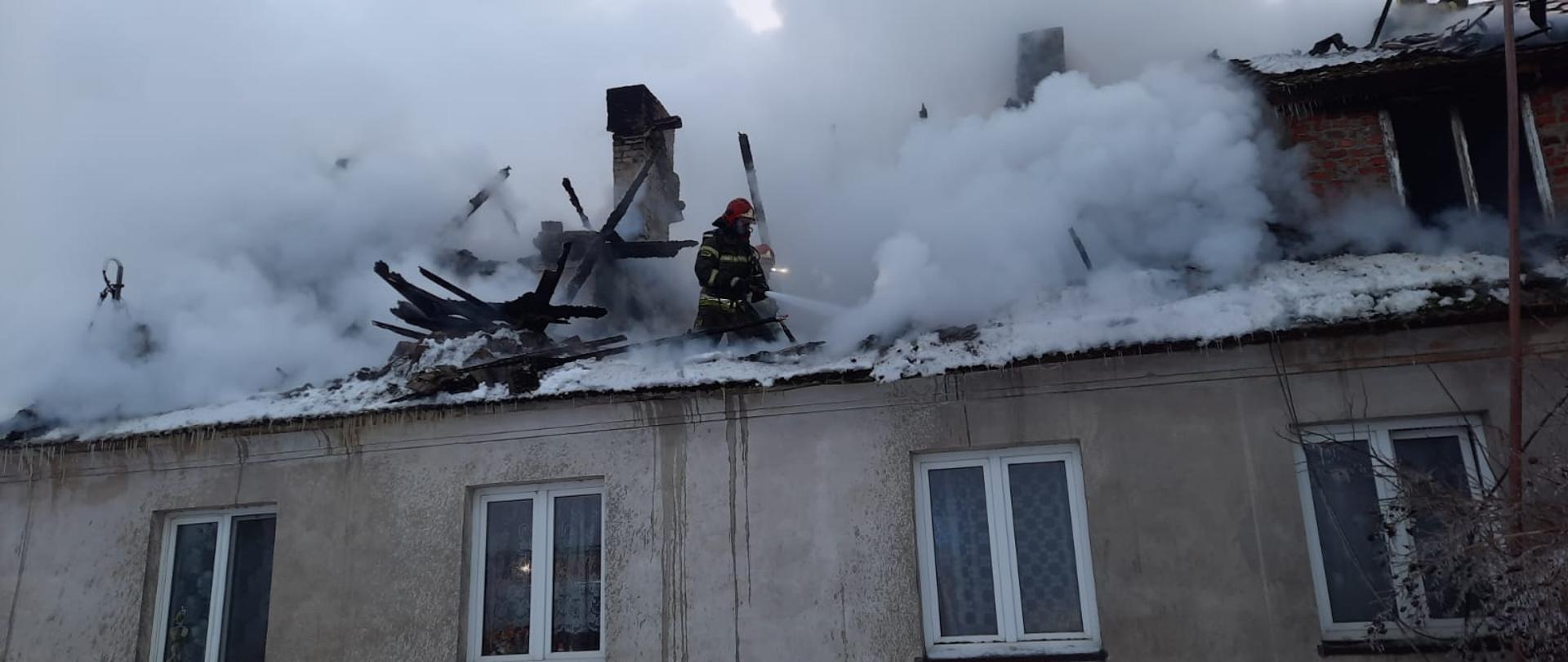 Pożar dachu i poddasza budynku mieszkalnego ze strażakami gaszącymi płomienie wodą stojąc na poddaszu przy dużym zadymieniu