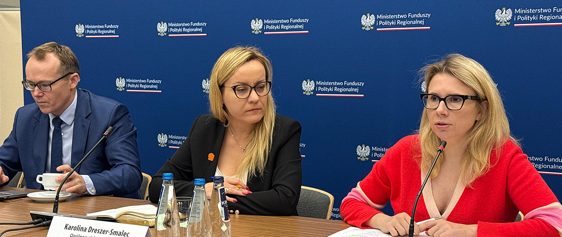 Trzy osoby siedzą przy stole konferencyjnym podczas spotkania w Ministerstwie Funduszy i Polityki Regionalnej. Za nimi znajduje się niebieska ścianka z logo i nazwą ministerstwa. Kobieta po prawej stronie mówi do mikrofonu, pozostałe osoby słuchają.