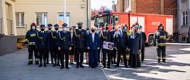 Fotografia przedstawia zdjęcie grupowe zaproszonych gości, a także strażaków, którzy ubrani są w ubrania specjalne. Za grupą znajduje się wóz strażacki, masz flagowy oraz budynek komendy.