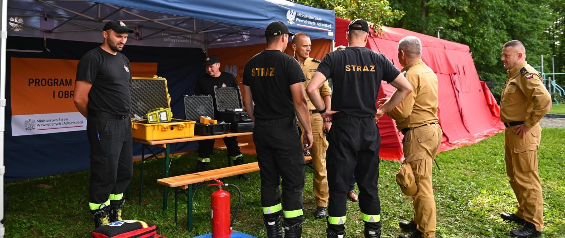 Przed namiotem oznakowanym napisami Ochrona Ludności stoją zebrani ludzie. Strażacy ubrani w czarne ubranie koszarowe oraz pisakowe mundury przygotowują stanowisko do prezentacji. Na przodzie namiotu rozłożony koc na którym leży manekin do ćwiczeń z resuscytacji krążeniowo oddechowej. Dookoła zielone drzewa.