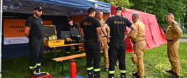 Przed namiotem oznakowanym napisami Ochrona Ludności stoją zebrani ludzie. Strażacy ubrani w czarne ubranie koszarowe oraz pisakowe mundury przygotowują stanowisko do prezentacji. Na przodzie namiotu rozłożony koc na którym leży manekin do ćwiczeń z resuscytacji krążeniowo oddechowej. Dookoła zielone drzewa.