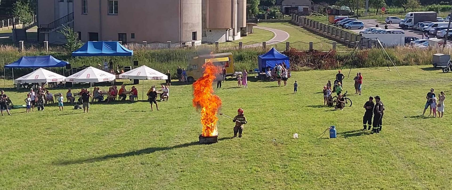 Widok na pokaz strażacki w którym strażak dolewa wody do płonącego oleju pokazując zjawisko wyrzutu. Na terenie pokazu widzowie oraz goście pikniku.
