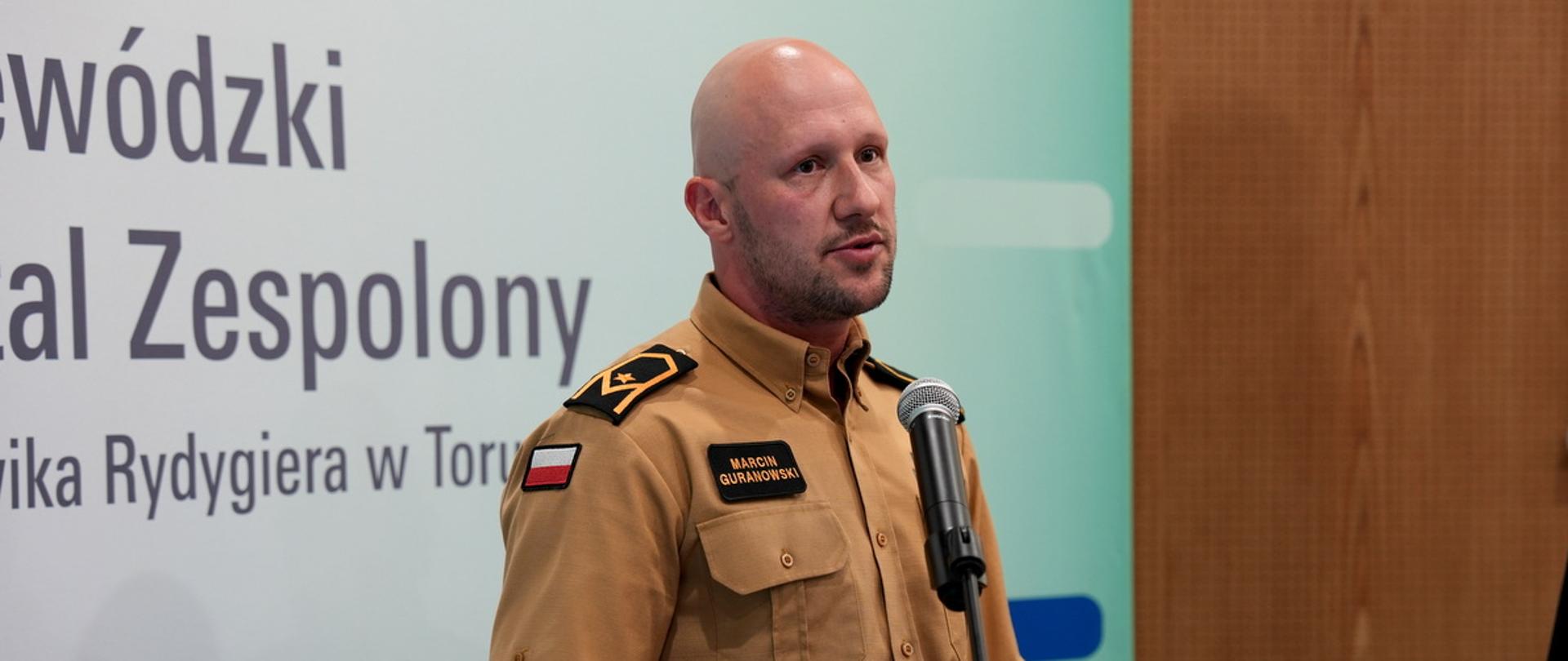 Portret z profilu strażaka w piaskowym mundurze z plakietką „Marcin Guranowski”, który mówi do mikrofonu podczas konferencji w szpitalu w Toruniu.