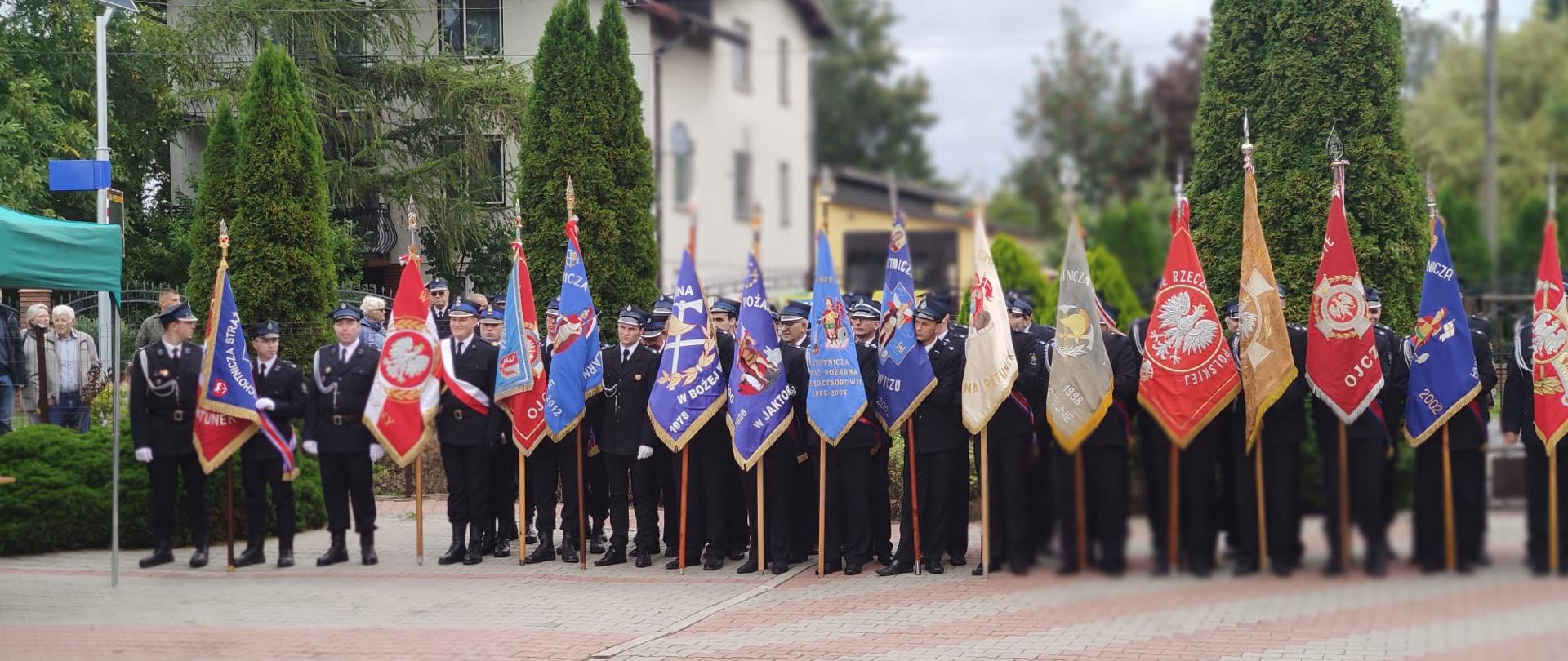 Strażacy w mundyrach galowych stoją ze sztandarami swoich jednostek w szeregu. W tle budynki.