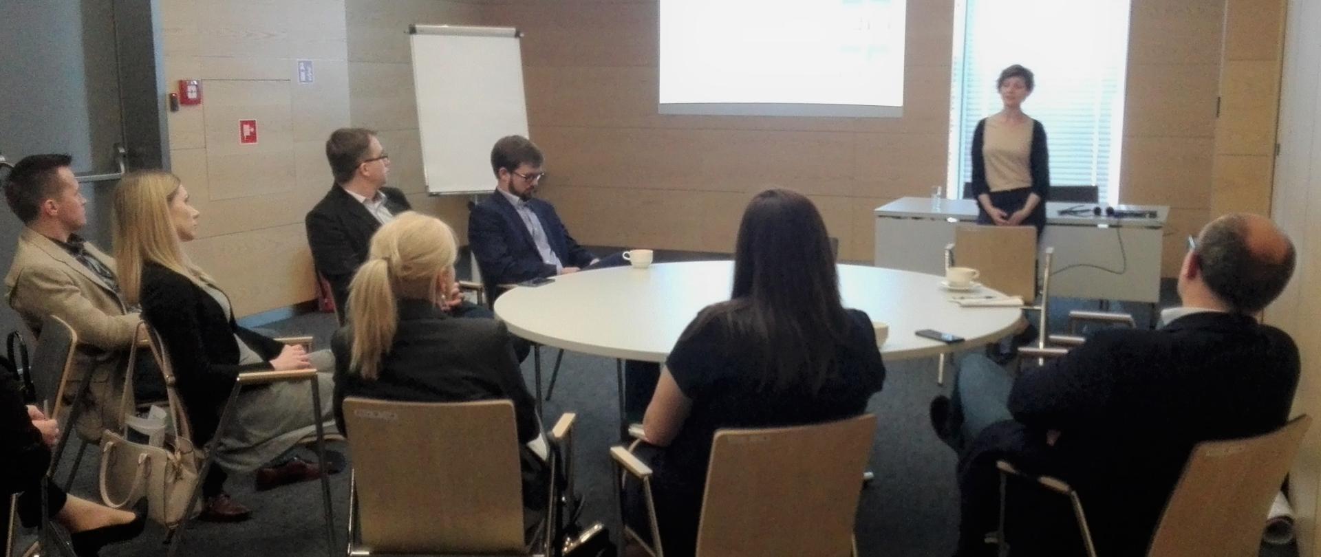 Dyrektor Agata Miazga na stojąco przemawia do kilkorga osób siedzących przy stole.