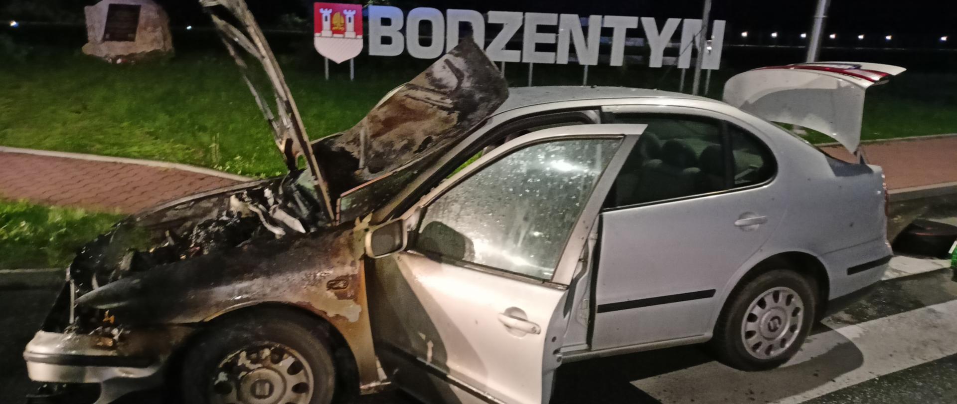 Zdjęcie przedstawia spalony samochód osobowy stojący na przejściu dla pieszych na tle ozdobnego napisu.