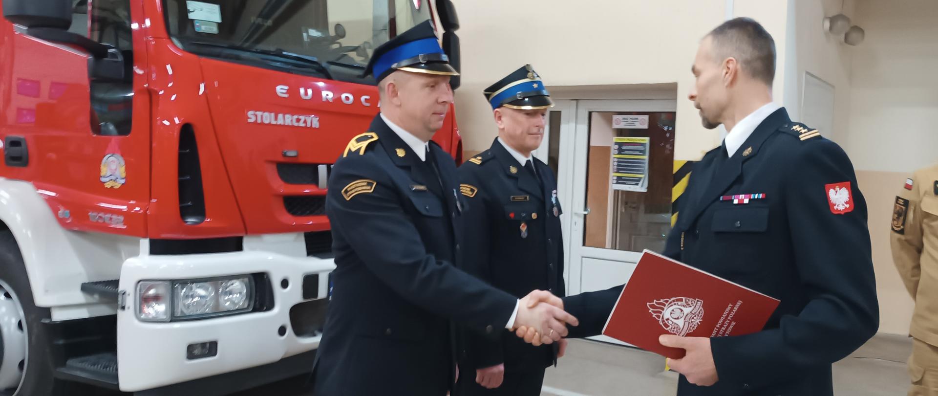 Odznaczeni strażacy otrzymują nagrodę od Komendanta Powiatowego Straży Pożarnej. W tle czerwony pojazd pożarniczy. 