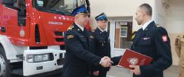 Odznaczeni strażacy otrzymują nagrodę od Komendanta Powiatowego Straży Pożarnej. W tle czerwony pojazd pożarniczy. 