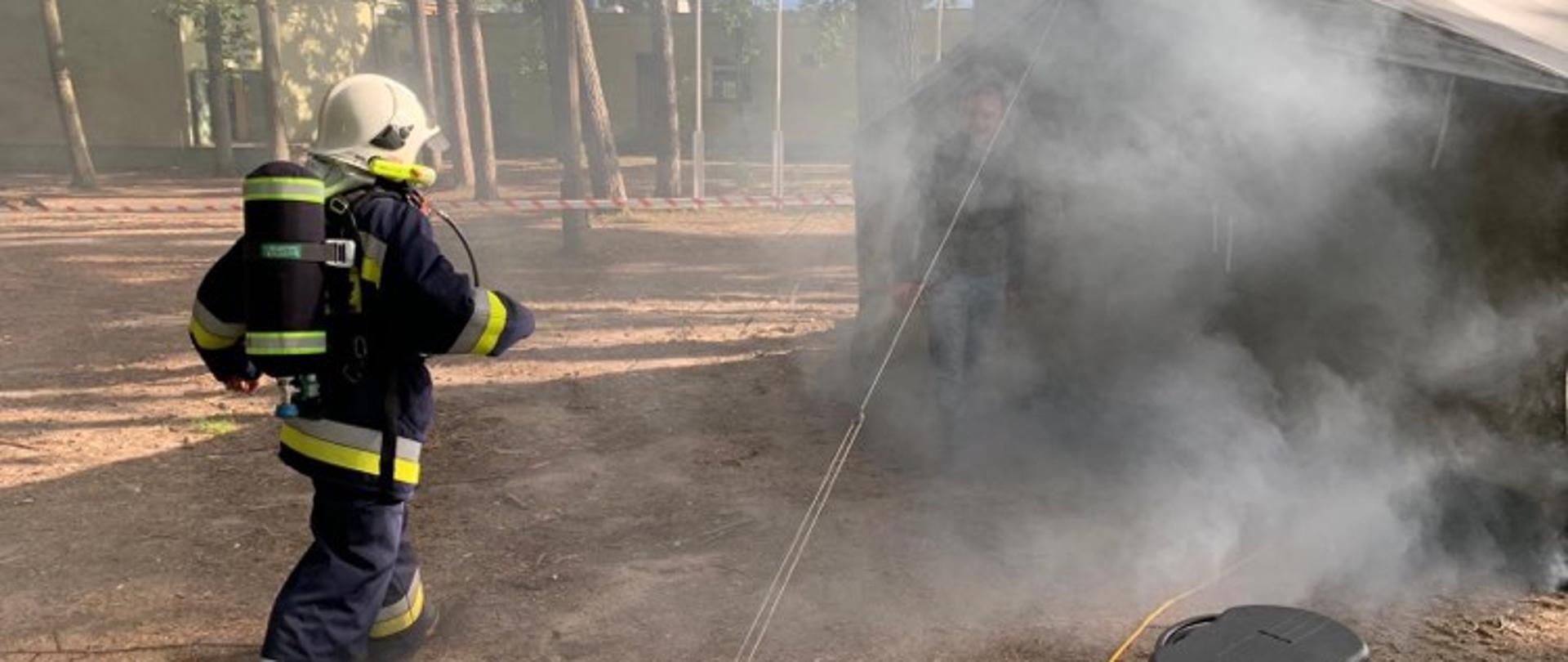 Zdjęcie przedstawia strażaka w umundurowaniu bojowym, który wchodzi do zadymionego namiotu