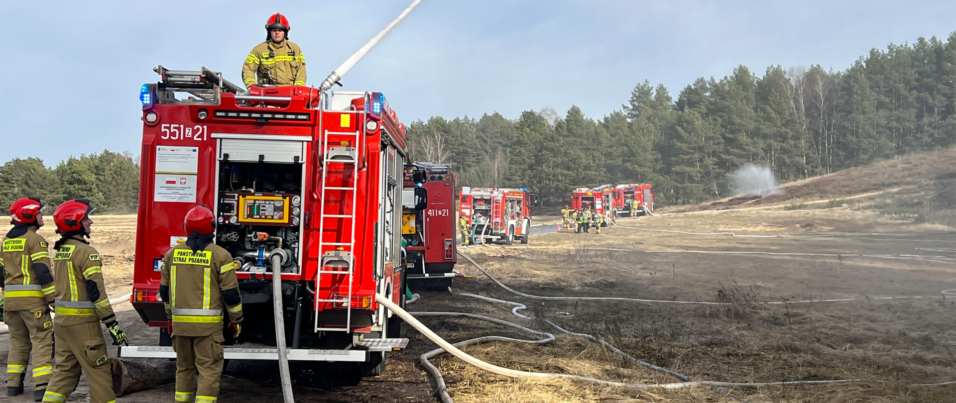 Pojazdy ratowniczo-gaśnicze, które stoją na drodze. Wokół samochodów widoczni są strażacy. W tle widać las. Zdjęcie powiększy się po kliknięciu na nie.